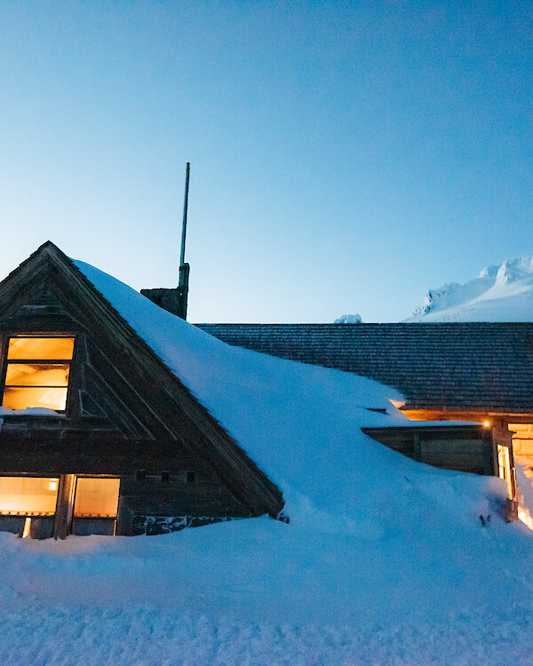 Silcox Hut pictured at dusk in the snow, with light streaming out of the windows and Mt. Hood looming in the background