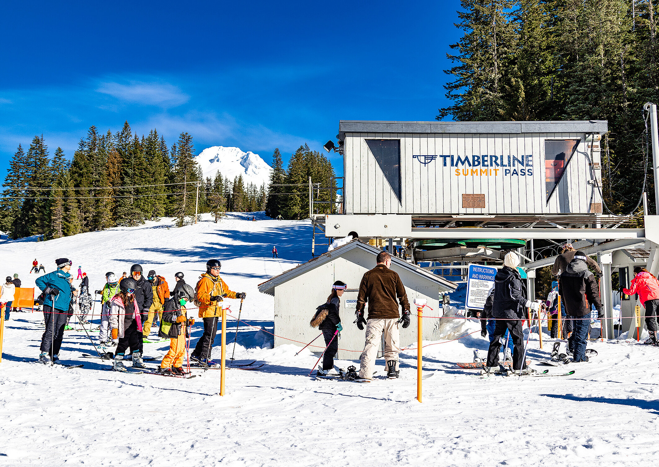 Skiers and snowboarders approaching the timberline summit pass chair lift on a blue bird day with snowy Mt. Hood in the background