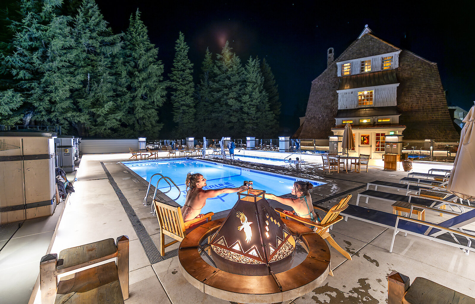 Two women clinking their beers together in Cheers by the fireplace at the Timberline outdoor swimming pool.