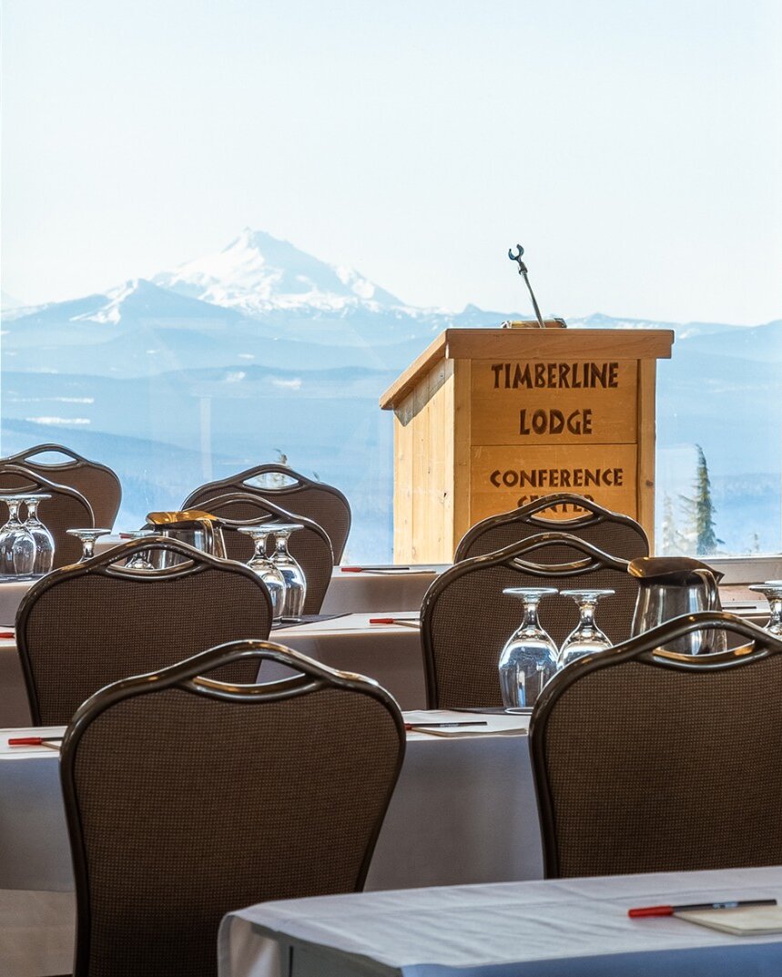Wooden podium with "Timberline Lodge Conference Center" carved into its front pictured in front of a window featuring views of Mt. Jefferson, facing tables and chairs set up for am eeting