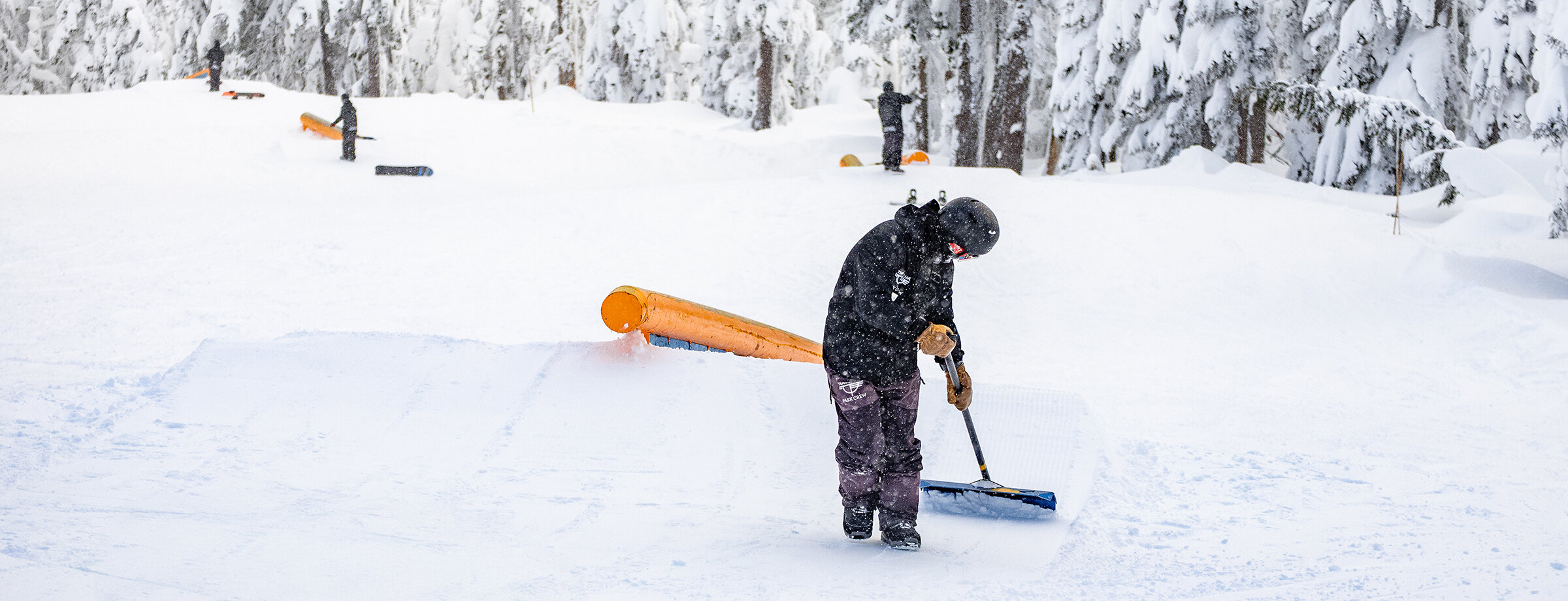 Timberline terrain park staff shaping a snow ramp around an orange rail feature during active snowfall, surrounded by snow-covered trees on Mount Hood.