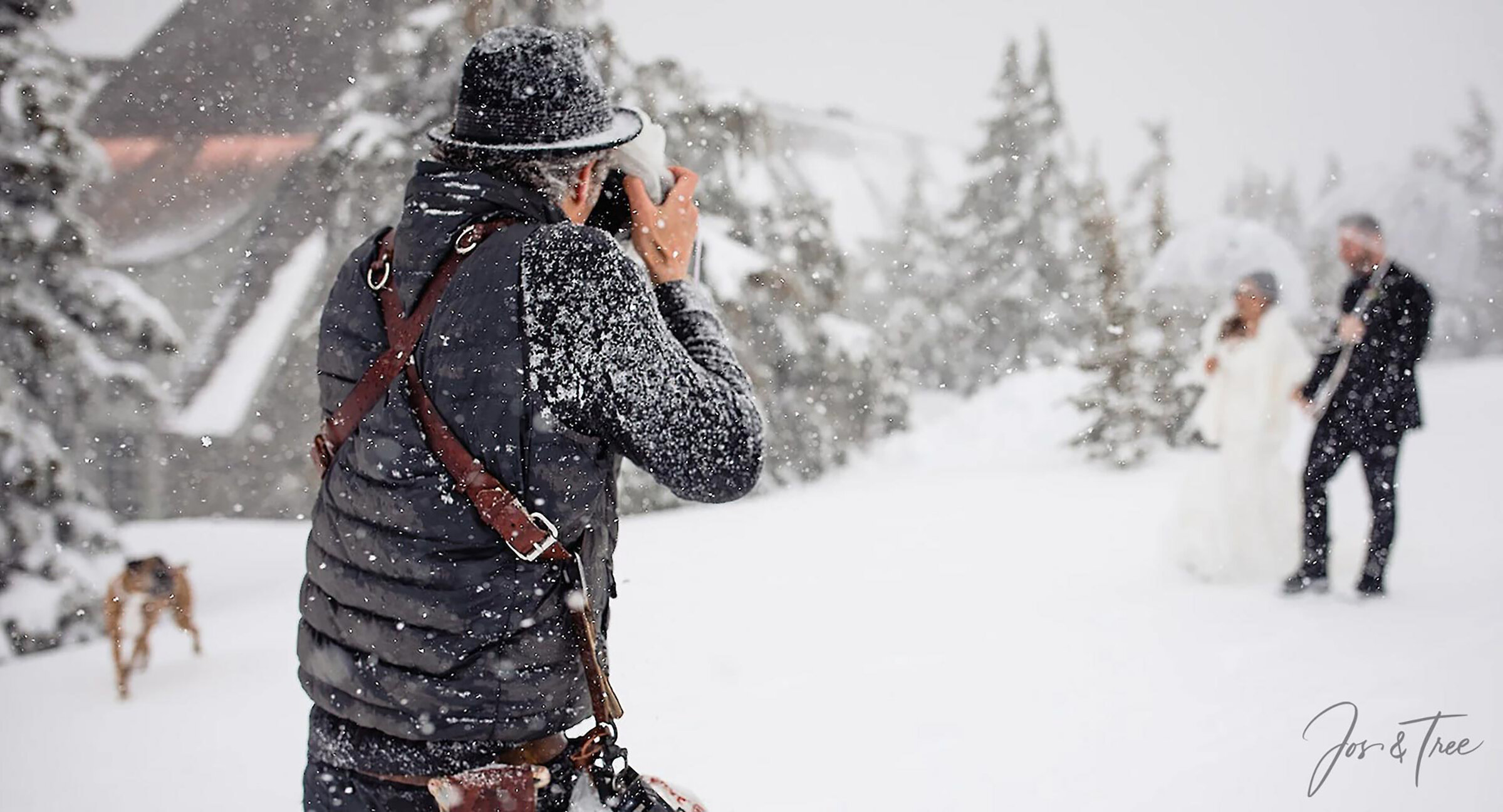 A photographer in winter gear taking photos in falling snow outside Timberline Lodge, with a bride and groom under clear umbrellas among snow-covered trees in the background.