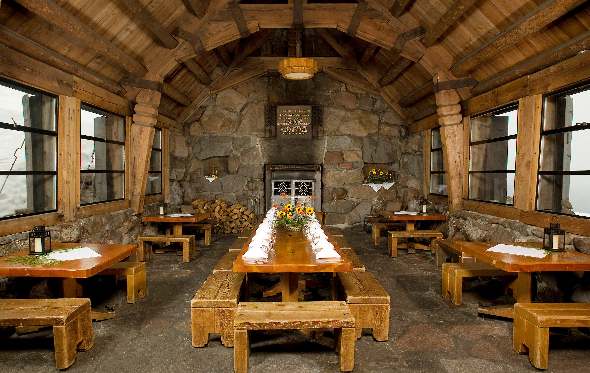 Rustic stone and timber room at Timberline Lodge set with long tables and benches for an event, with stacked firewood and warm lighting.